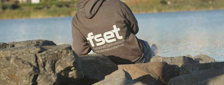 FSR-Mitglied von hinten am Ufer sitzend, Pullover ist hinten mit Logo bedruckt