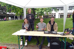 Drei Studierende sitzen auf einer Bank, zwei davon hinter einer Kasse und einer hinter einem DJ-Mischpult