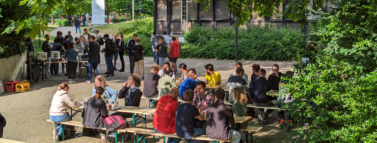 Studierende sitzen an Tischen in einem Innenhof und unterhalten sich