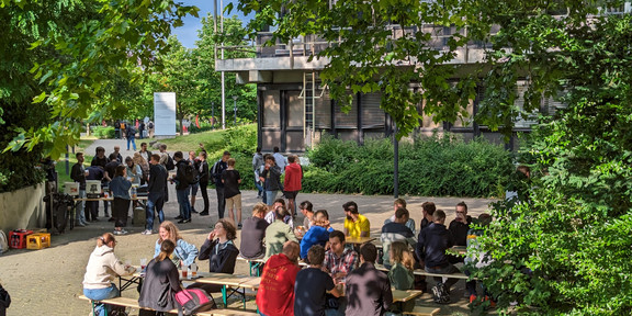 Studierende sitzen an Tischen in einem Innenhof und unterhalten sich