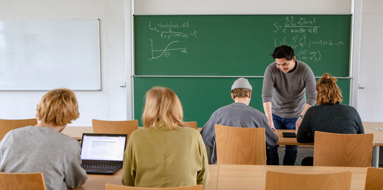 Vier Studierende sitzen im Seminarraum zur Tafel gewandt. Der Dozierende erklärt etwas.