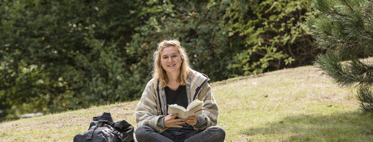 Eine Studentin sitzt im Schneidersitz auf einer Wiese mit einem Buch in der Hand.