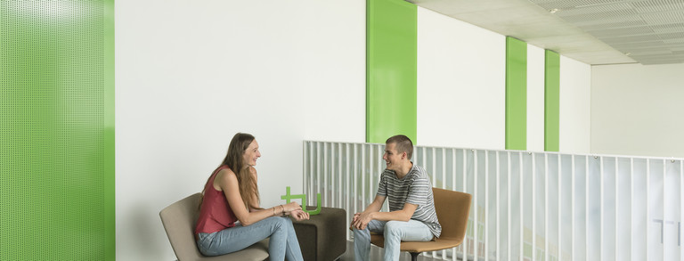 Zwei Studierende sitzen auf Stühlen an einem Tisch, auf dem das TU Dortmund Logo steht. Hinter ihnen befindet sich ein Handlauf und eine Treppe.