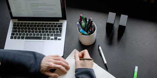 A student working at a table with a laptop, pencils and a notebook.